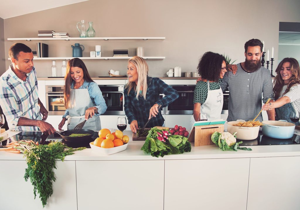 Beispielbild für Küchen Prreusser, Küchenstudio Bad Homburg. Sechs Freunde (zwei Männer und vier Frauen) die glücklich sind kochen gemeinsam und bereiten Essen vor auf einer weissen Kücheninsel. Zwei Frauen haben Kochschürzen an. Darauf befindet sich Spargel, Gemüse,Orangen, Karotten, Tomaten, Weingläser mt Rotwein. Pasta wird in einem Topf gekocht. Im Hintergrund befinden sich Küchengeräte sowie weisse Regale mit Bücher und Deko und Kerzen darauf. Die Wand ist beige oder hellbraun.