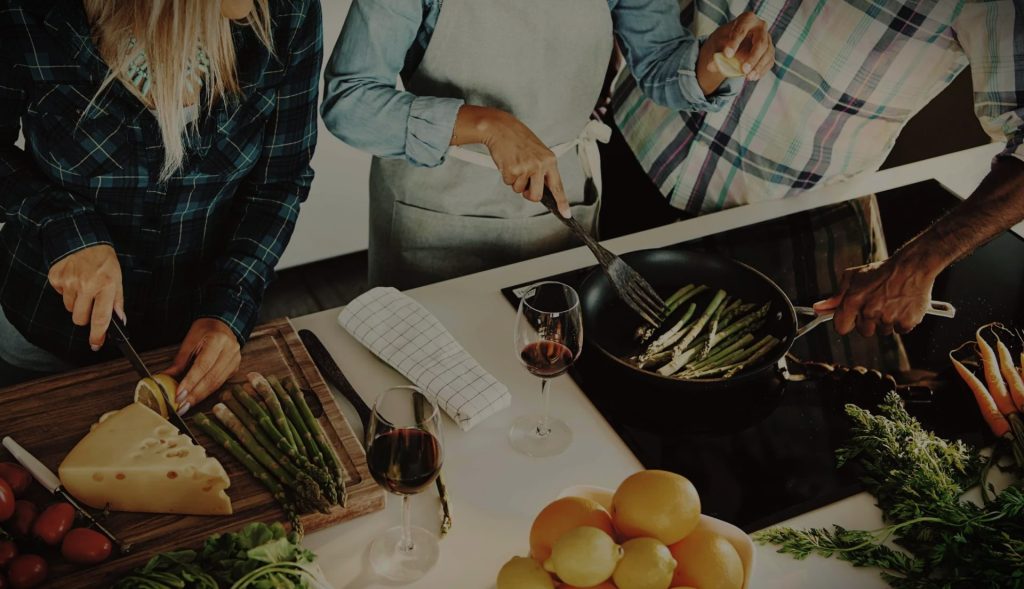 Beispielbild für Küchen Preusser - Küchenstudio Hanau. Drei Menschen kochen gerade auf der Küchenzeile, wo sich auch ein Herd befindet und darauf eine Pfanne mit grünem Spargel darin. Die Pfanne halet ein Mann in kariertem Hemd und den Spargel dreht eine Frau mit Küchenschürze mit einem Pfannenwender/Küchenutensil und presst mit der anderen Hand eine halbe Zitrone aus auf den Spargel darauf. Auf der Kücheninsel befindet sich auch eine Schüssel mit Orangen und Zitronen. Die dritte Frau in kariertem Hemd schneidet gerade eine Zitrone auf einem Holzbrett mit einem Messer. Auf dem Holzbrett befinden sich auch noch grüner Spargel, Käse und Tomaten. Zwei Weingläser mit Rotwein stehen auch auf der Küchenzeile und ein Geschirrtuch.