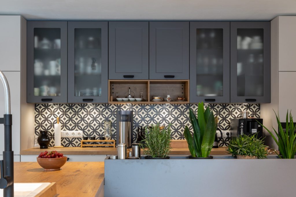 Beispielbild für Küchen Preusser - Küchenstudio Kefenrod. Auf dem Bild erkennt man eine Kücheninsel mit Holzobefläche wo man einen dunkelgrauen Wasserhahn und eine Holzschale mit roten Weintrauben sieht. Daneben steht eine Art tiefe Trennwand aus Beton wo grüne Pflanzen darin sind als Abtrennung zurr Einbauküche in grau matt. Die Einbauküche hat geschlossene Schranktüren sowie Milchglas Türen wo man das Geschirr bzw. Gläser darin erkennen kann. Auch zwei kleine Holzauschnitte sind in der grauen Küchenwand mit weißem Geschirr darin. Man kann eine schwarze Kaffeemaschine, einen Wasserkocher und eine Küchenrolle sehen auf der Küchenzeile. Und zwischen Küchenzeile und Küchenschränken oben ist die Wand mit gemusterten Fließen als Hintergrund.