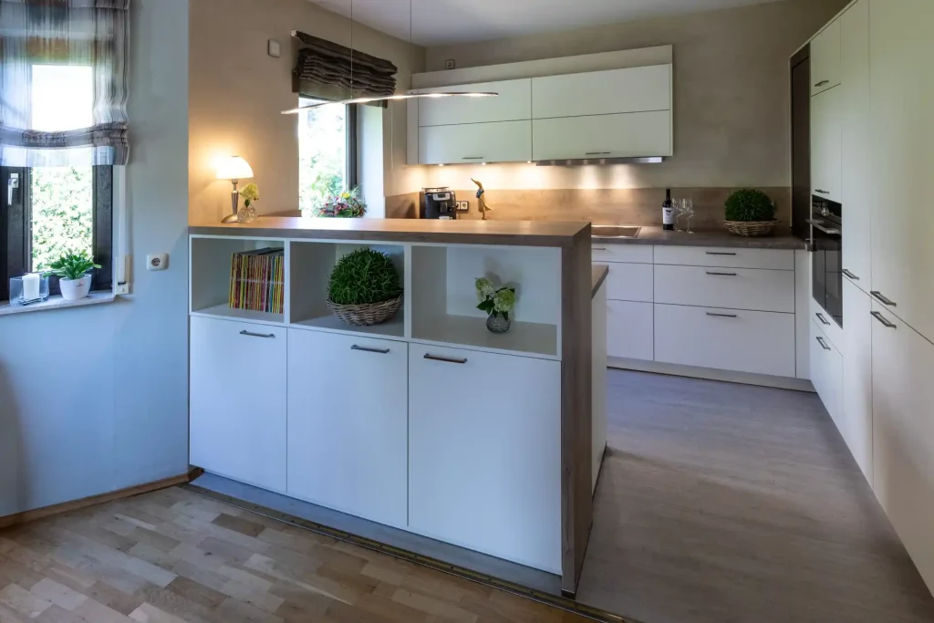 Beispielbild für Küchen Preusser - Küchenstudio Altenstadt. Ein Holzboden ist zu sehen und eine weisse Kpche mit hellen Holzelementen. Ein fenster ist zu sehen mit einem bunten Vorhang darüber. Eine lange Lampe hängt von der Decke. Grüne Pflanzen stehen auf der Fensterbank sowie in einem weissen Schrank und auf der Küchenfläche sowie eine Weinflasche und zwei Weingläser.