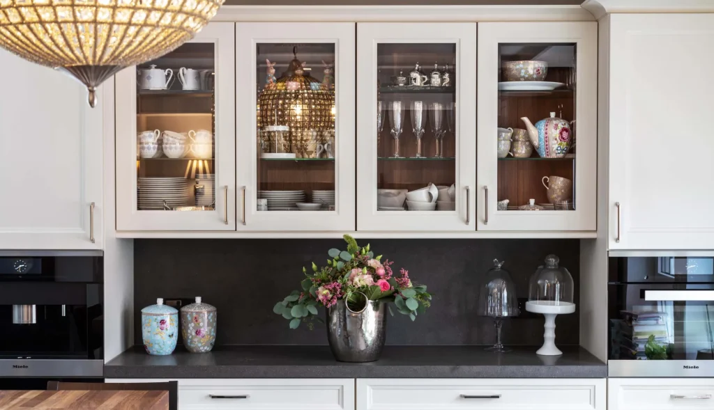 Beispielbild für Küchen Preusser - Küchenstudio Bad Soden-Salmünster. Weisse Einbauschränke sind im Hintergrund mit einer Vitrine voll mit Geschirr und Gläsern. Eine Kaffeemaschine ist auch zu sehen auf der grauen Ablagefläche und bunte Blumen in einer silbernen Vase.