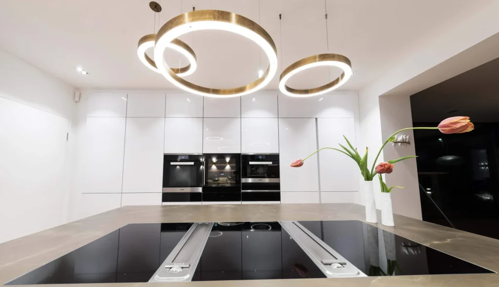 Beispielbild für Küchen Preusser - Küchenstudio Bad Vilbel, wo sich eine helle Küche befindet. Drei kreisförmige Lampen hängen von der Decke. Die Einbauküche besteht aus weissem Hochglanz. Orange Tulpen in einer weissen Vase stehen auf der Kücheninsel.