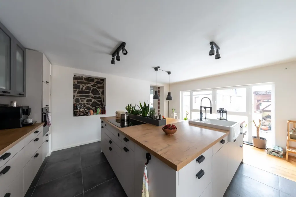 Beispielbild für Küchen Preusser - Küchenstudio Brachttal, wo sich eine weisse Küche mit Holzablagen befindet. Dunkelgraue Lampen hängen von der Decke herunter. Eine Kaffeemaschine ist zu sehen und Pflanzen auf der Kücheninsel die aus Holz ist. Eine Schüssel Weintrauben steht auch darauf. Fenster bzw. Glastüren sind in der hellen weissen Küche auch zu sehen. Die Küche hat einen dunkelgrauen Steinboden und beim Fenster befindet sich ein Holzboden. In de weissen Wand befindet sich ein ausgeschnittenes Steinfenster.