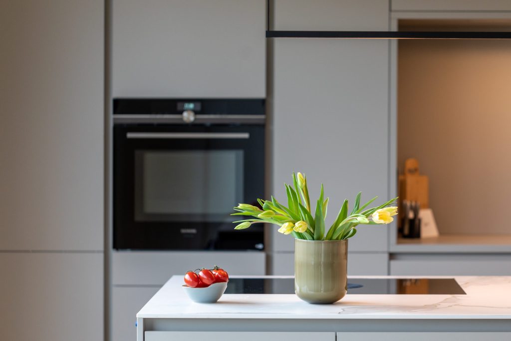 Beispielbild für Küchen Preusser - Küchenstudio Bruchköbel, wo sich eine helle graue Küche befindet. Man kann auf dem Bild den eingebauten Ofen in schwarz sehen und auf der hellgrauen Kücheninsel sind gelbe Tulpen in einer Vase zu sehen und Tomaten in einer Schüssel.