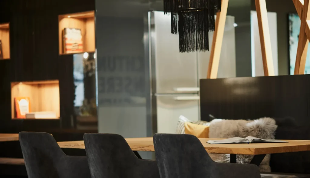 Beispielbild für Küchen Preusser - Küchenstudio Frankfurt, wo sich ein Holztisch mit dunkelgrünen Stühlen aus Stoff befinden. Ein offenes Buch liegt am Tisch und im Hintergrund sind flauschige Kissen zu sehen. Es ist eine angenehme Atmosphäre mit warmen Licht überall. Eine schwarze Lampe hängt von der Decke.