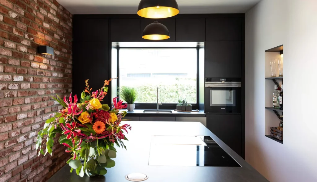Beispielbild für Küchen Preusser, Küchenstudio Gedern. Auf dem Bild ist eine große, dunkelgraue Kücheninsel zu sehen und darüber hängen zwei grau, orange Leuchten und auf der Kücheninsel befinden sich bunte Blumen in gelb, orange und pink. Links daneben ist eine dunkelrote Ziegelwand mit einer grauen Leuchte. Rechts ist eine weiße Wand mit einem Ausschnitt wo Weingläser und Weinflaschen darin sind. Ganz dahinter befindet sich ein großes Fenster mit Aussicht ins grüne und ein anderes Gebäude kann man auch sehen. Vor dem Fenster befindet sich ein Wasserhahn, daneben grüne Pflanzen.
