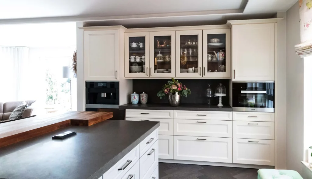 Beispielbild für Küchen Preusser - Küchenstudio Kronberg. Weisse Einbauschränke sind im Hintergrund mit einer Vitrine voll mit Geschirr und Gläsern. Eine Kaffeemaschine ist auch zu sehen auf der grauen Ablagefläche und bunte Blumen in einer silbernen Vase. Und auch noch mehr Geschirr auf der dunkelgrauen Küchenzeile bzw. Küchenoberfläche. Rechts befindet sich eine kleine grüne Stoffbank. Und links befindet sich eine beige Couch aus Leder mit einem Kissen darauf und dahinter eine Glasfront wo davor eine Stehlampe in grau steht.