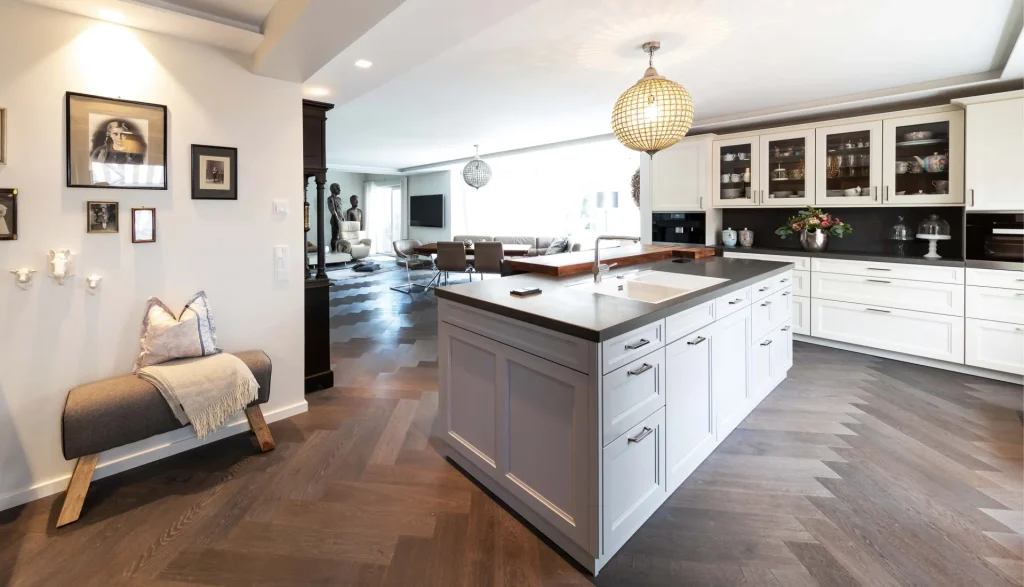 Beispielbild für Küchen Preusser, Küchenstudio Nidda. Auf dem Bild befindet sich eine große aus weißem matten Holz befindliche Kücheninsel auf einem dunkelbraunen Parkett-Boden. Bei der Kücheninsel sieht man einen Wasserhahn und das Waschbecken. Die Oberfläche der Kücheninsel ist dunkelgrau. Eine runde Lampe (Luster) hängt von der Decke genau über der Kücheninsel. Rechts daneben befindet sich eine Einbauküche, auch weiss und matt mit geschlossenen Türen sowie Glasfronten, wo man Geschirr darin sieht. Links neben der Kücheninsel befinden sich auf der Wand Bilder in verschiedenen Größen (Portraits) und darunter steht eine kleine Stoffbank in grau und mit einem hellen Kissen und einer hellen Decke darauf. Ganz hinten im Raum sieht man noch einen grossen braunen Küchentisch mit Lederstühlen rundherum. Und dahinter sieht man noch einen ganz bequemen einzelnen Couchstuhl bzw. Chefsessel mit Statuen/Skulpturen dahinter und einem Flachbildschirm (Fernseher) auf der Wand hängend.