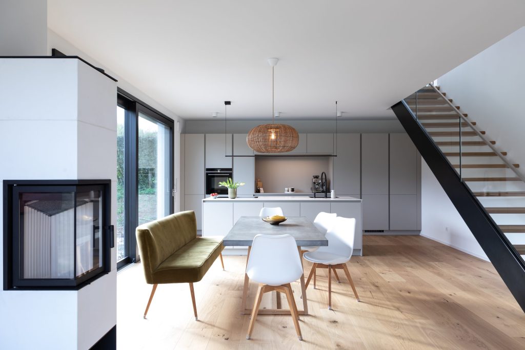 Beispielbild für Küchen Preusser - Küchenstudio Rosbach. Eine hellgraue Einbauküche befindet sich am Bild und ein hellgrauer Küchentisch auf einem Holzboden befindet Weisse Stühle und eine grüne Samtbank steht beim Küchentisch.. Eine Schale Obst mit Bananen und Äpfel steht am Tisch. Und auf der Kücheninsel vor der Einbauküche steht eine Vase mit gelben Tulpen. Man kann auf dem Bild das Waschbecken und die Wasserleitung sehen und eine Kaffeemaschine. Eine Art schwebende dunkle Treppe ist rechts zu sehen. Eine Deckenleuchte in braun hängt auch von der Decke genau über dem Küchentisch. Auch ein heller (Holz-) Ofen ist iim Zimmer zu sehen und Glastüren die zum Garten hinaus führen.